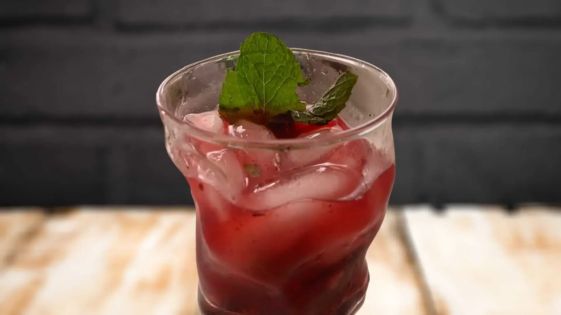 A refreshing red drink with ice and mint leaves, served in a curved glass against a dark background, representing a Bourbon Blackberry Smash.