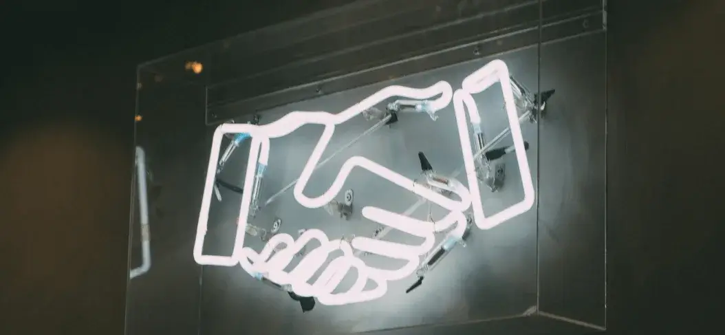 A glowing neon sign of two hands shaking, symbolizing agreement and partnership, against a dark background.