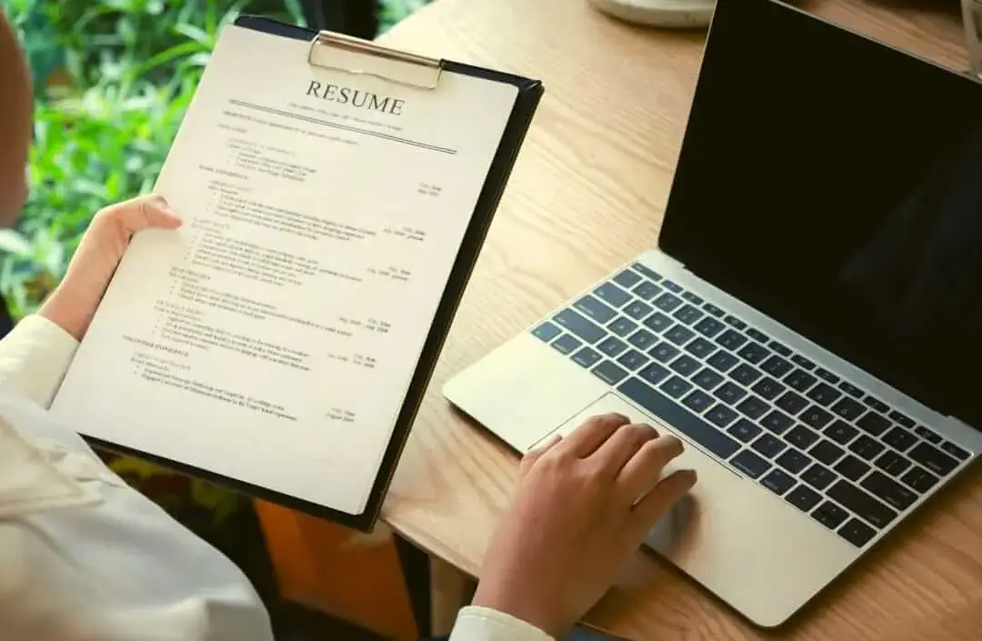 Person reviewing a printed resume on clipboard while using a laptop on a wooden desk.