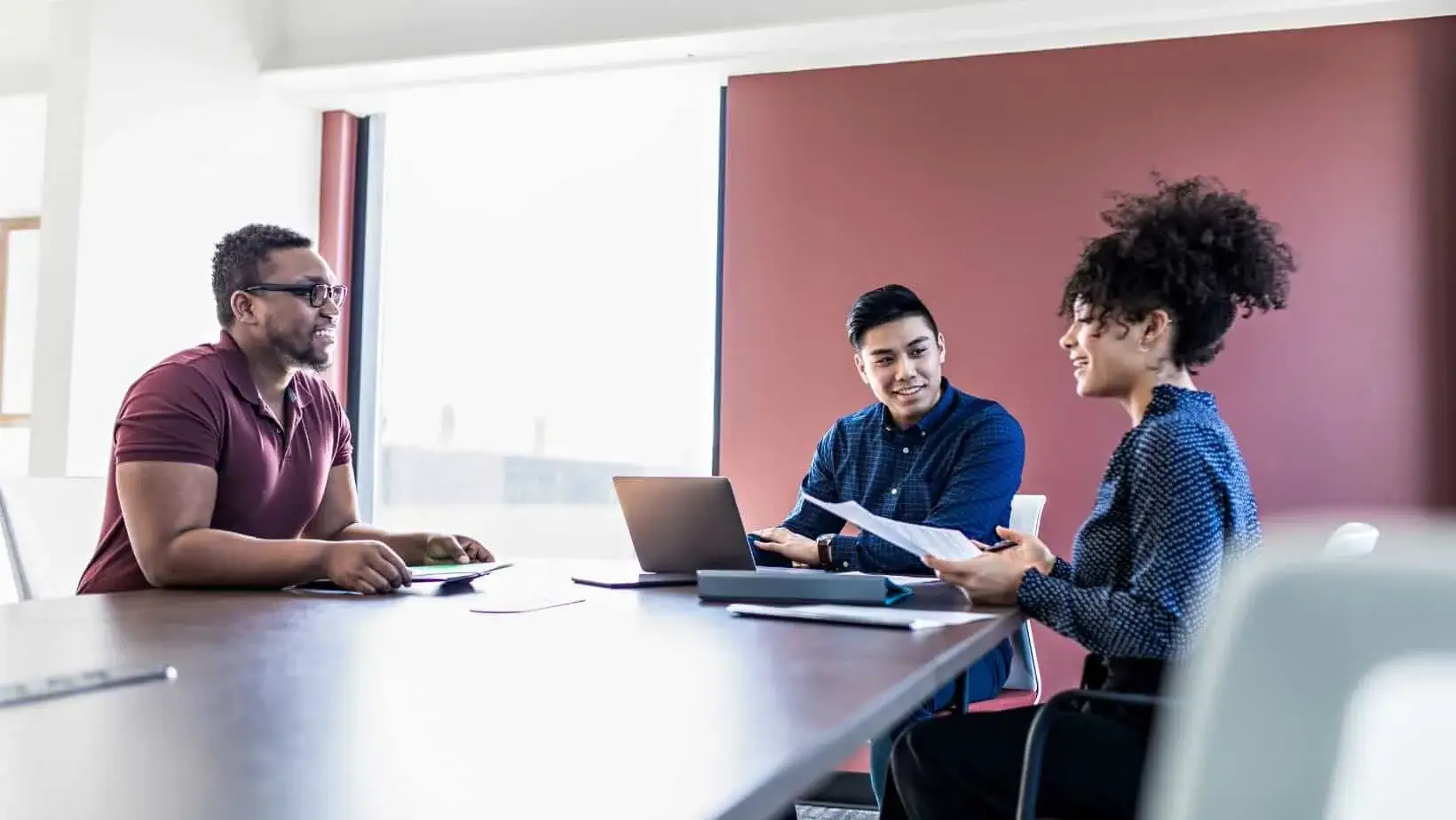 A diverse group of professionals engaged in a collaborative discussion around a conference table, reviewing documents and using a laptop.