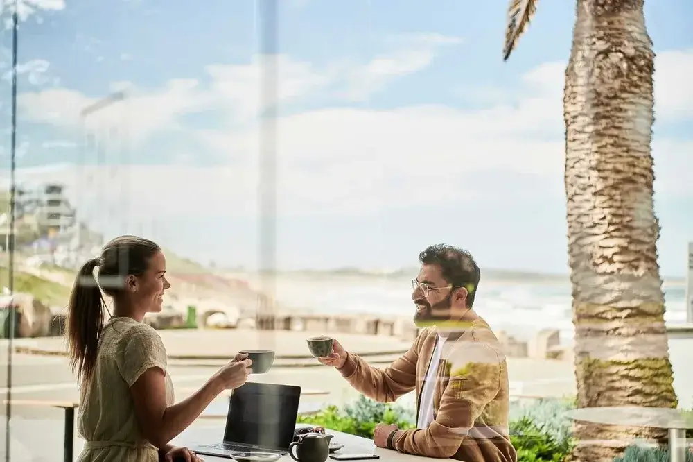 A woman and a man enjoying coffee at a seaside café with palm trees and ocean views, and a laptop on the table.