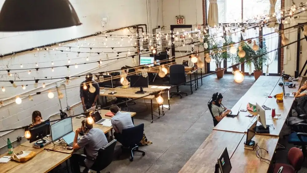 Open office space with people working at desks under hanging string lights.