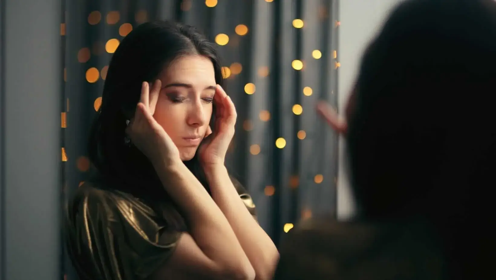 A woman stands in front of a mirror with her eyes closed and fingers on her temples, expressing signs of stress or burnout