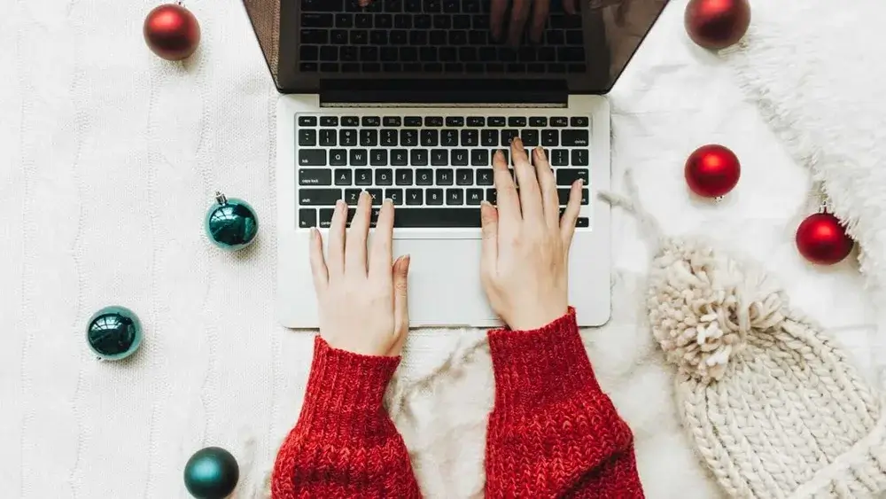 Person's hands typing on a laptop surrounded by festive ornaments and a cozy knit hat on a textured blanket.