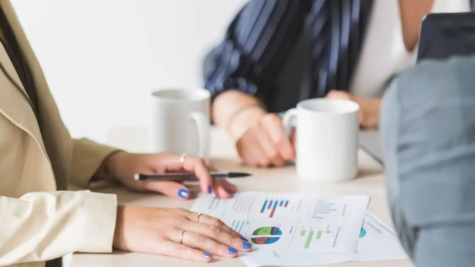Two professionals discuss documents with graphs and charts on a table, accompanied by coffee mugs and a laptop in a bright, modern workspace.