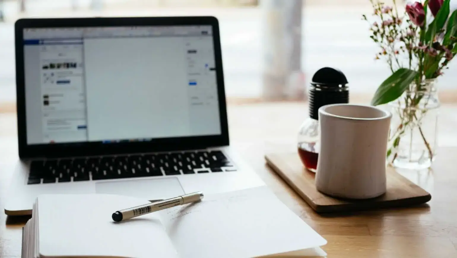 A cozy workspace setup with a laptop, an open notebook with a pen, a coffee mug, and a vase of fresh flowers on a wooden desk.