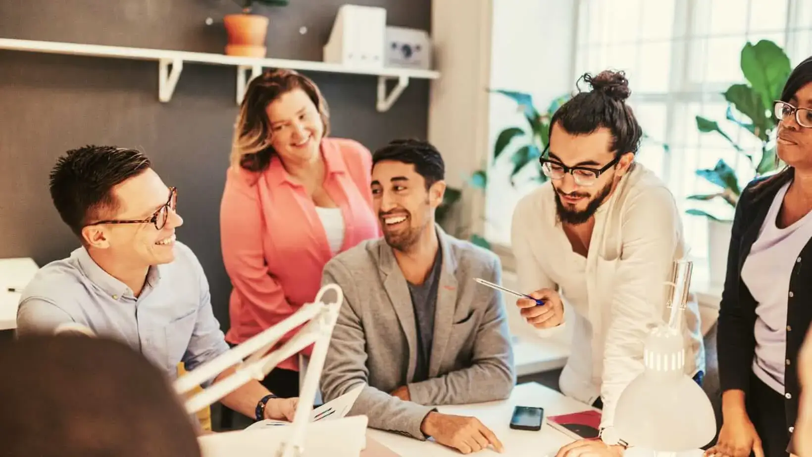 A group of five people engaged in a collaborative work session around a desk, surrounded by plants and office supplies.