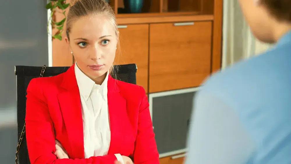 Woman in a red blazer with arms crossed, engaged in a serious conversation.