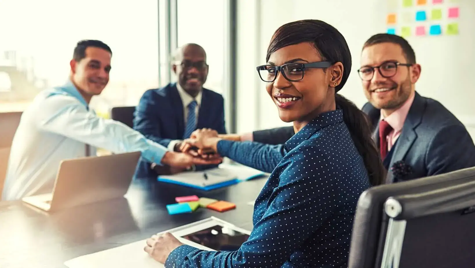 Smiling business professionals sitting around a table stack their hands in unity, showing teamwork and leadership.