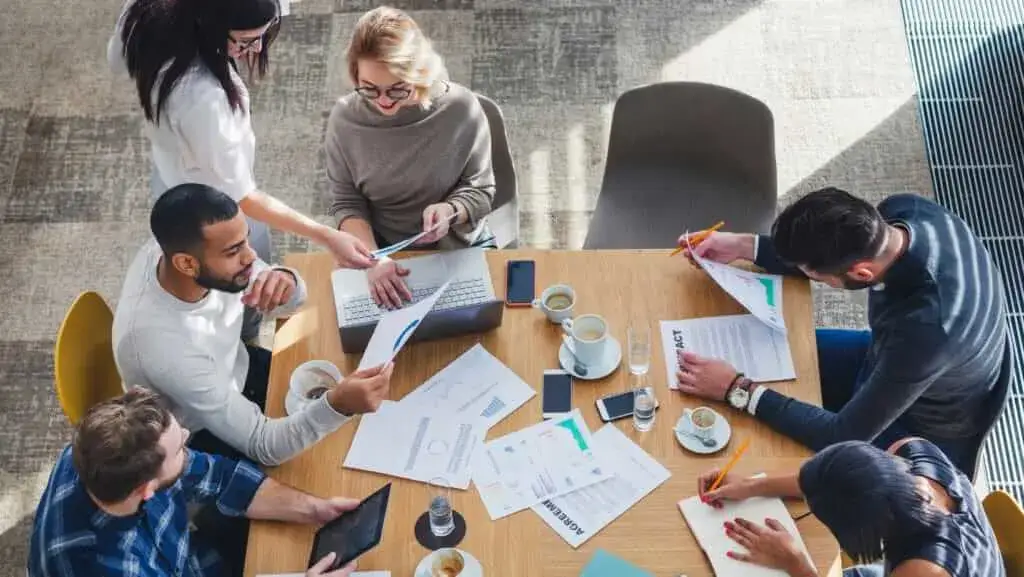 A group of people collaborates around a table, reviewing documents, laptops, and cups of coffee in a bright, modern workspace, promoting creativity.