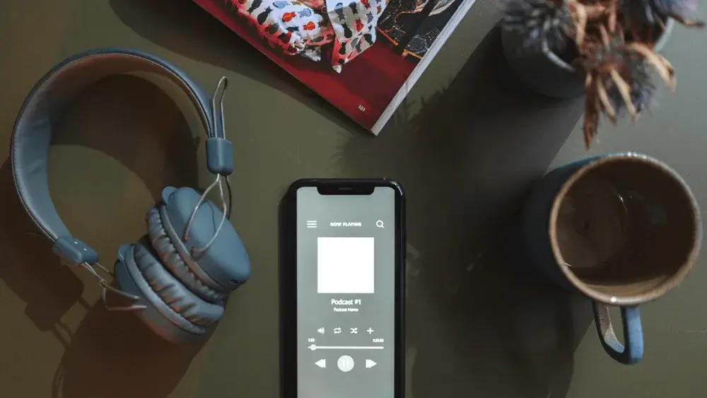 Flat lay of a smartphone playing a podcast, with headphones, coffee mug, and magazine on a table.