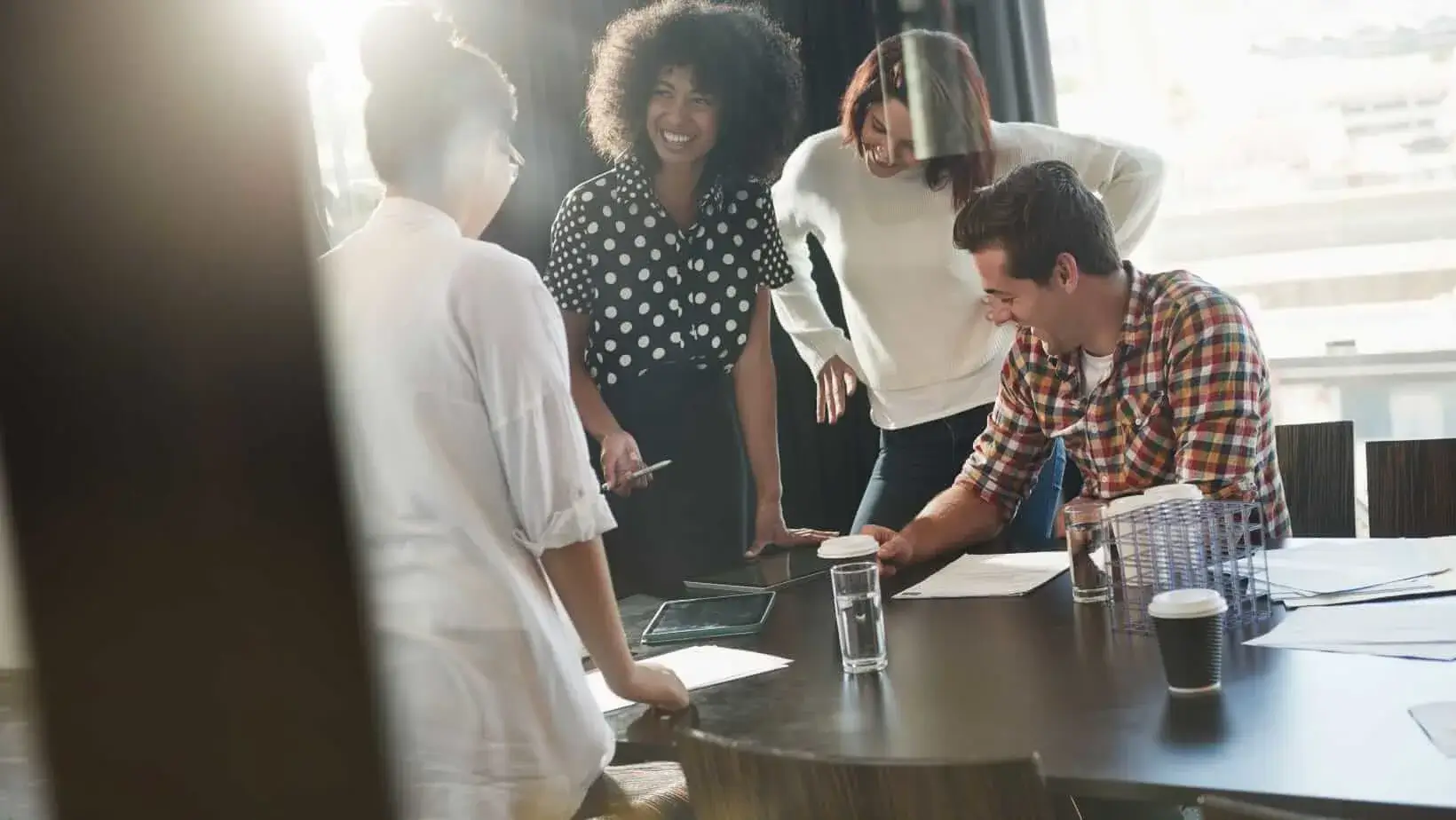 Coworkers smiling and collaborating around a table in a sunlit office meeting room.