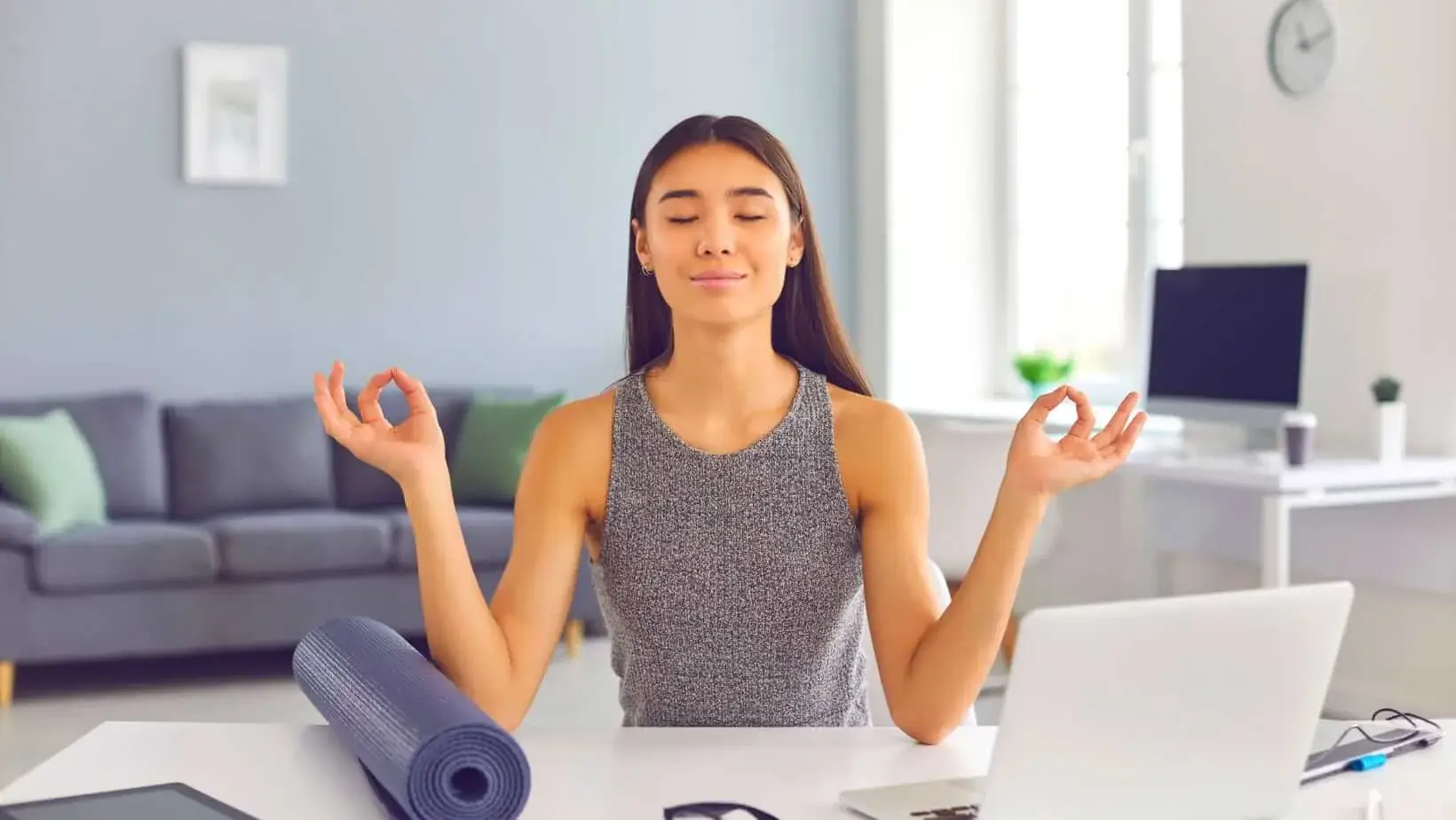 Woman meditating at her desk with eyes closed, a yoga mat beside her, and a laptop in front.