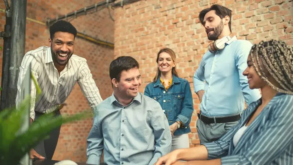Inclusive team of coworkers, including a person with Down syndrome, laughing and collaborating in a modern workspace.