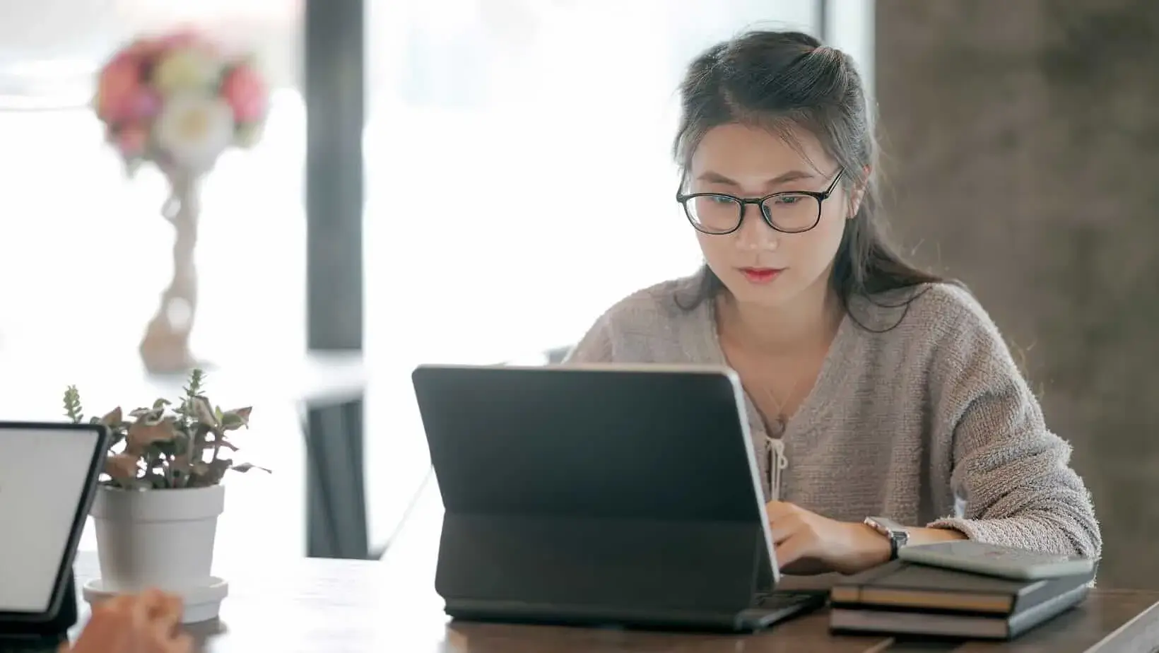 Woman wearing glasses focused on working at a laptop in a bright, modern workspace with books and a plant nearby.