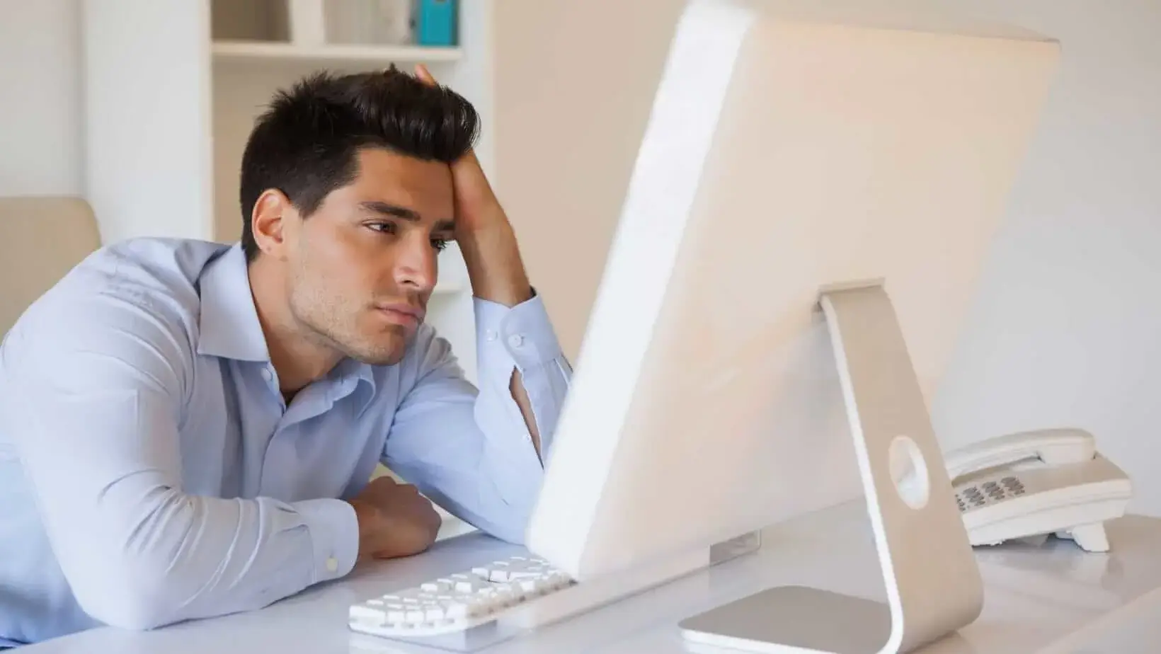 Frustrated office worker staring at a computer screen with a hand on his head.