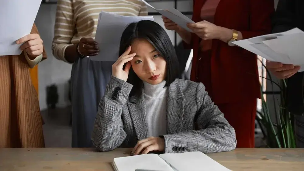 Stressed businesswoman surrounded by coworkers handing her documents, symbolizing workplace overwhelm