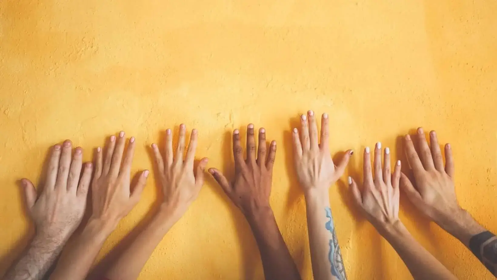 Diverse hands with different skin tones and nail designs against a yellow wall, symbolizing unity, diversity, and inclusion in the workspace.