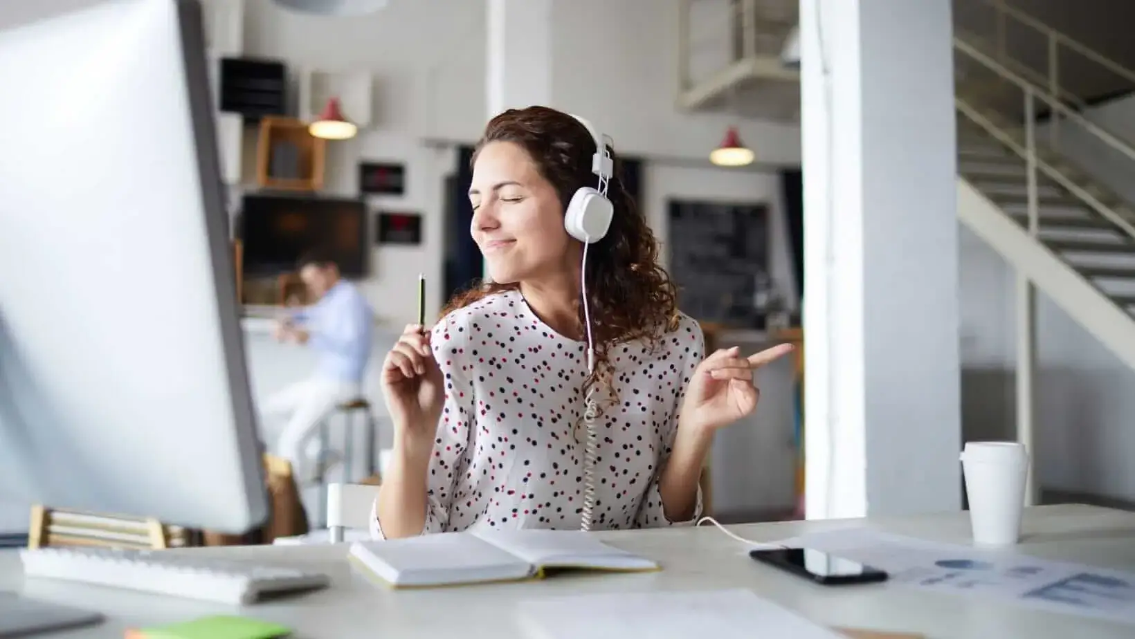 Woman wearing headphones, smiling and dancing while working at her desk, enjoying music during work.