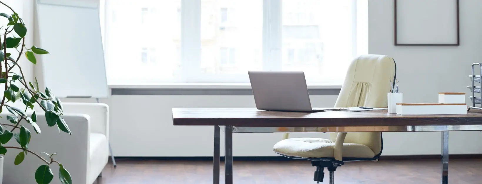 Modern minimalist office with a laptop on wooden desk, cream chair, indoor plant, and natural light from large windows.