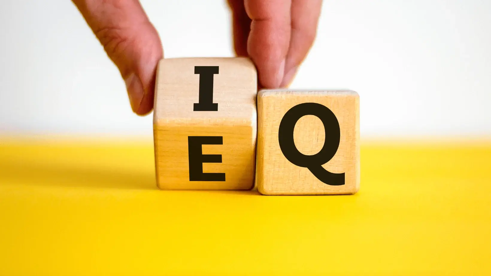 Wooden blocks displaying "IEQ" and "EQ" with a hand rotating the "I" block, symbolizing emotional vs. intellectual intelligence