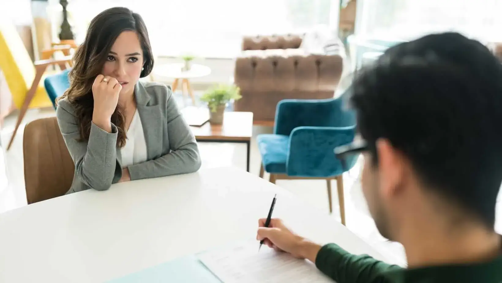 A woman in a grey blazer appears anxious during a job interview while a man holding a pen reviews her resume.