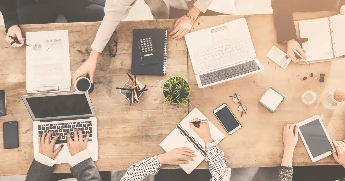 Aerial view of a collaborative workspace with hands writing, laptops, notebooks, and coffee cups on a wooden table.