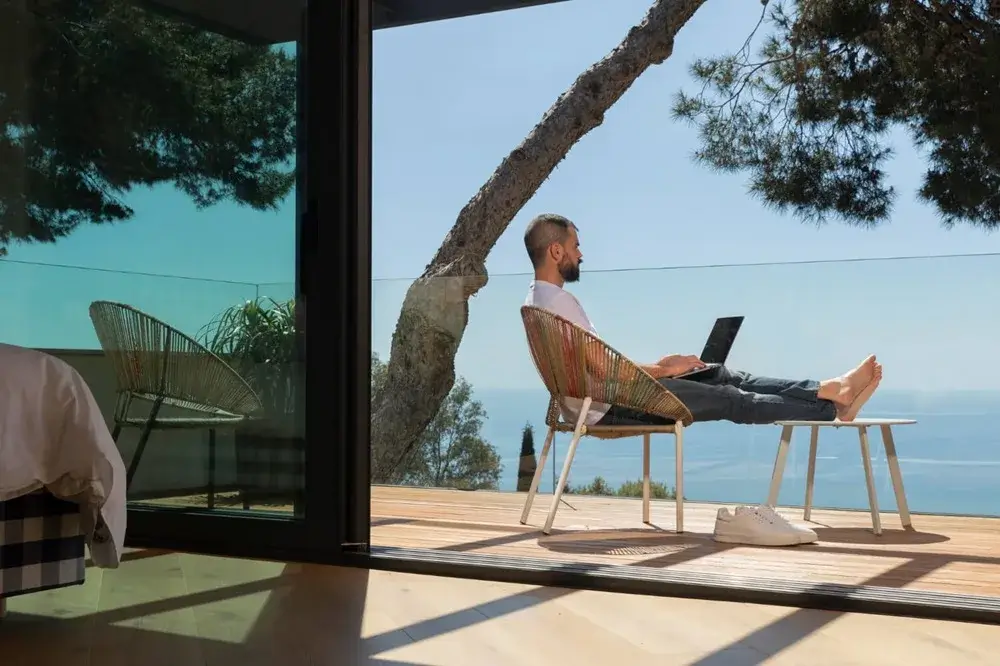 Person working on a laptop from a deck chair, overlooking the ocean and greenery, representing remote work in a scenic setting.