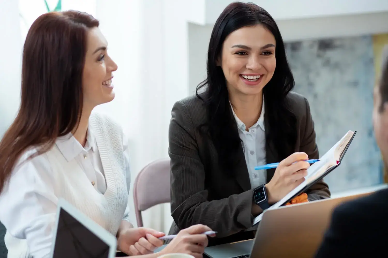 Two women smiling and talking during a business meeting with notebooks and laptops.