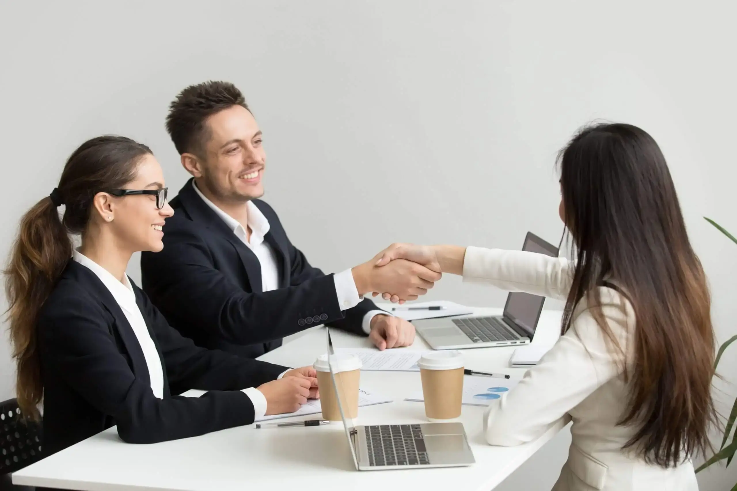 A woman in a white blazer shakes hands with a male interviewer while a female interviewer looks on, all smiling during a job interview.