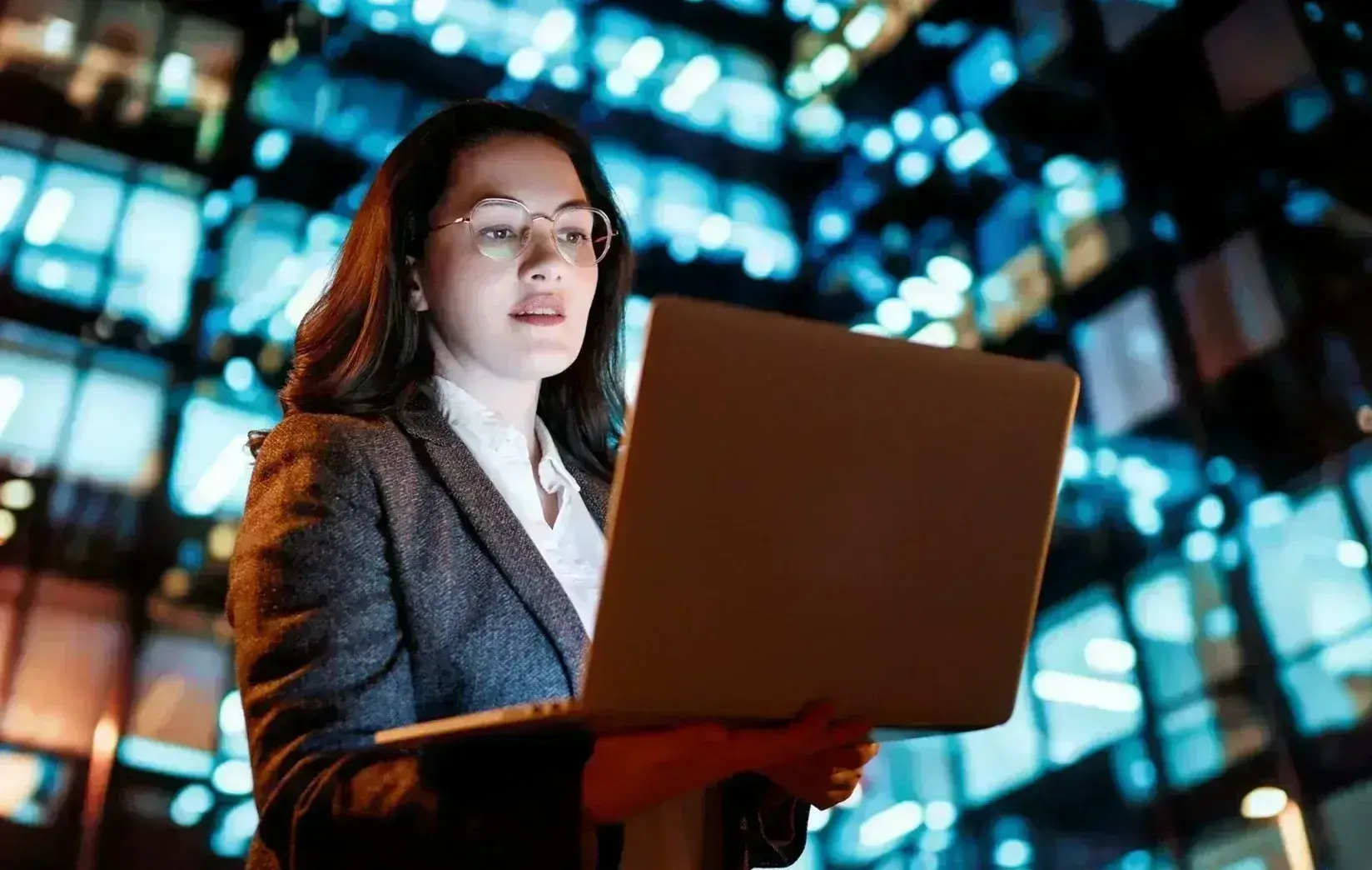 Businesswoman holding laptop at night in front of illuminated city building, suggesting technology and modern work culture.