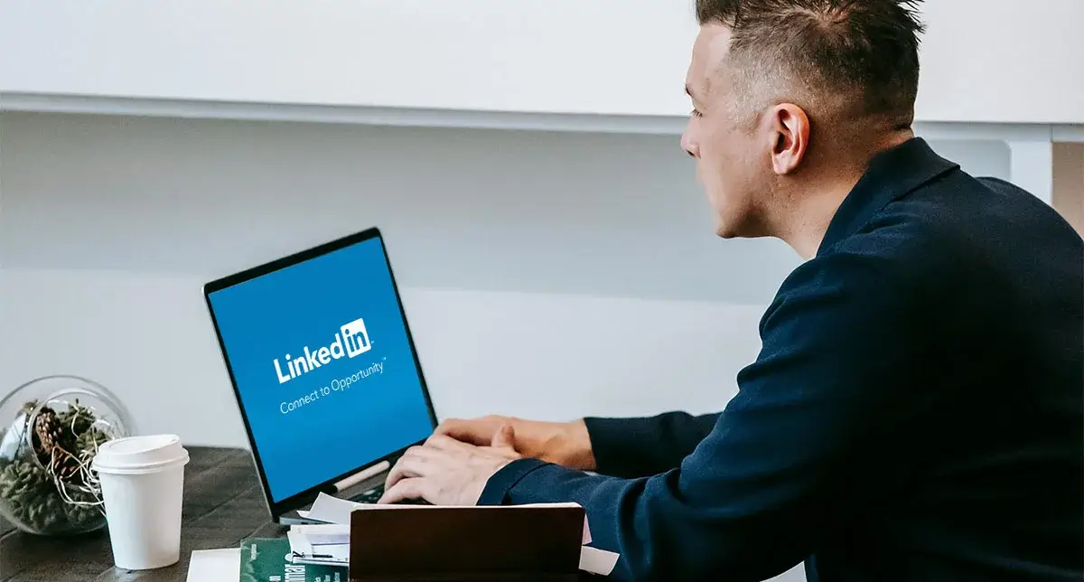 Man working on a laptop displaying the LinkedIn logo, seated at a desk with papers and a coffee cup nearby.