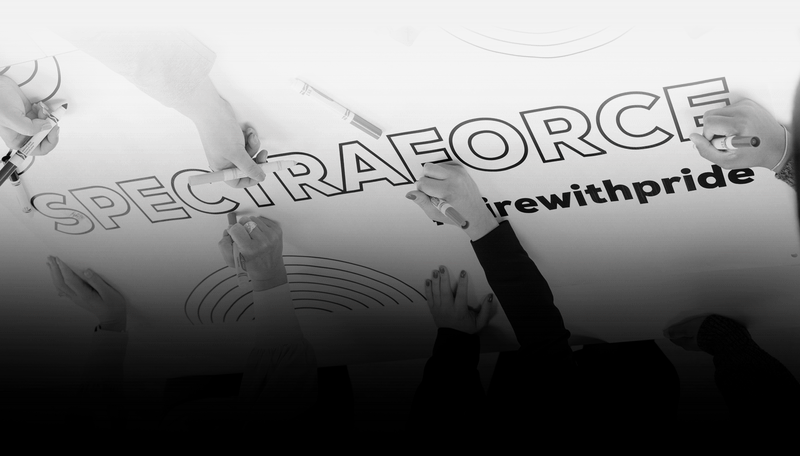 Hands holding markers, writing "SPECTRAFORCE #hirewithpride" on a large poster, showcasing teamwork and creativity.