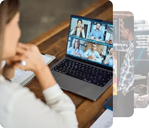 A woman sits at a wooden table, focused on her laptop screen showing multiple participants in a video call, with documents and a pen placed beside her.