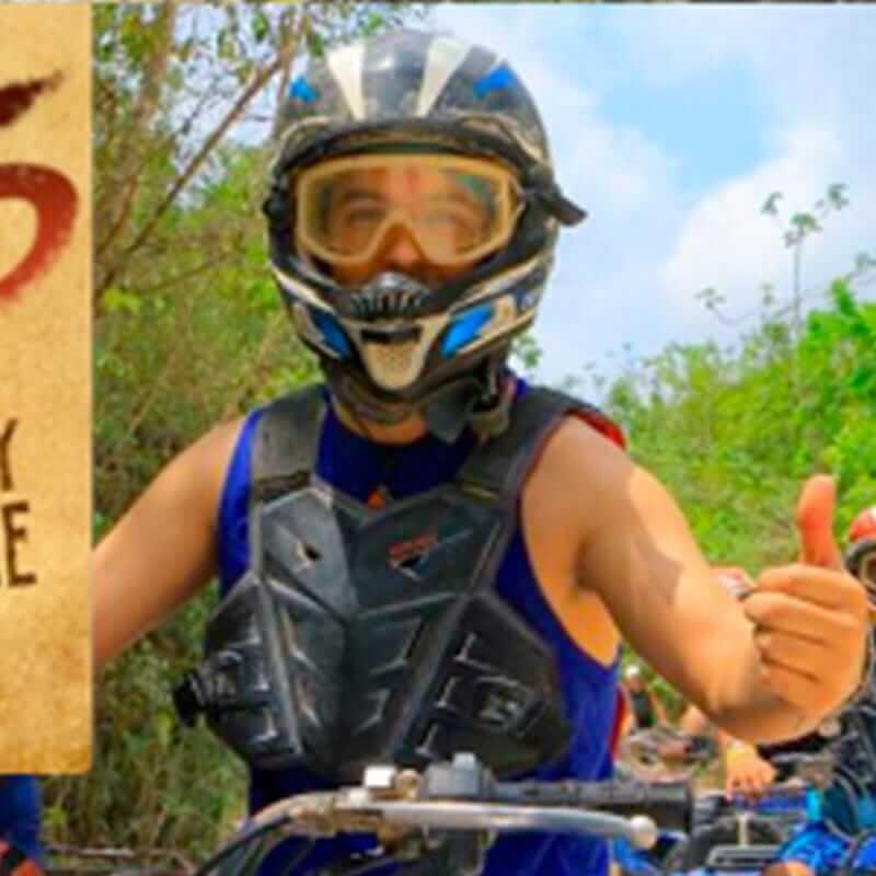 A person wearing a motocross helmet, goggles, and protective gear is riding an off-road vehicle. They are giving a thumbs-up sign with their left hand. The background features trees and a partly cloudy sky.