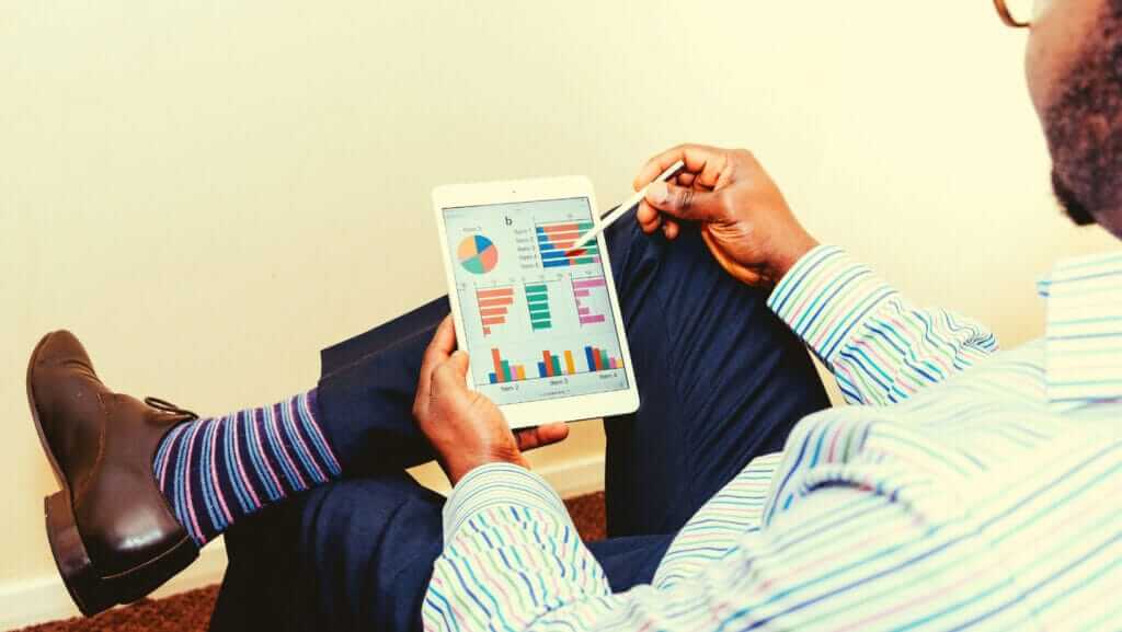 A person wearing a striped shirt, striped socks, and dress shoes sits cross-legged while using a stylus to interact with a tablet displaying colorful charts and graphs. The background is neutral, showcasing the focus required by inspirational women in their pursuit of excellence.