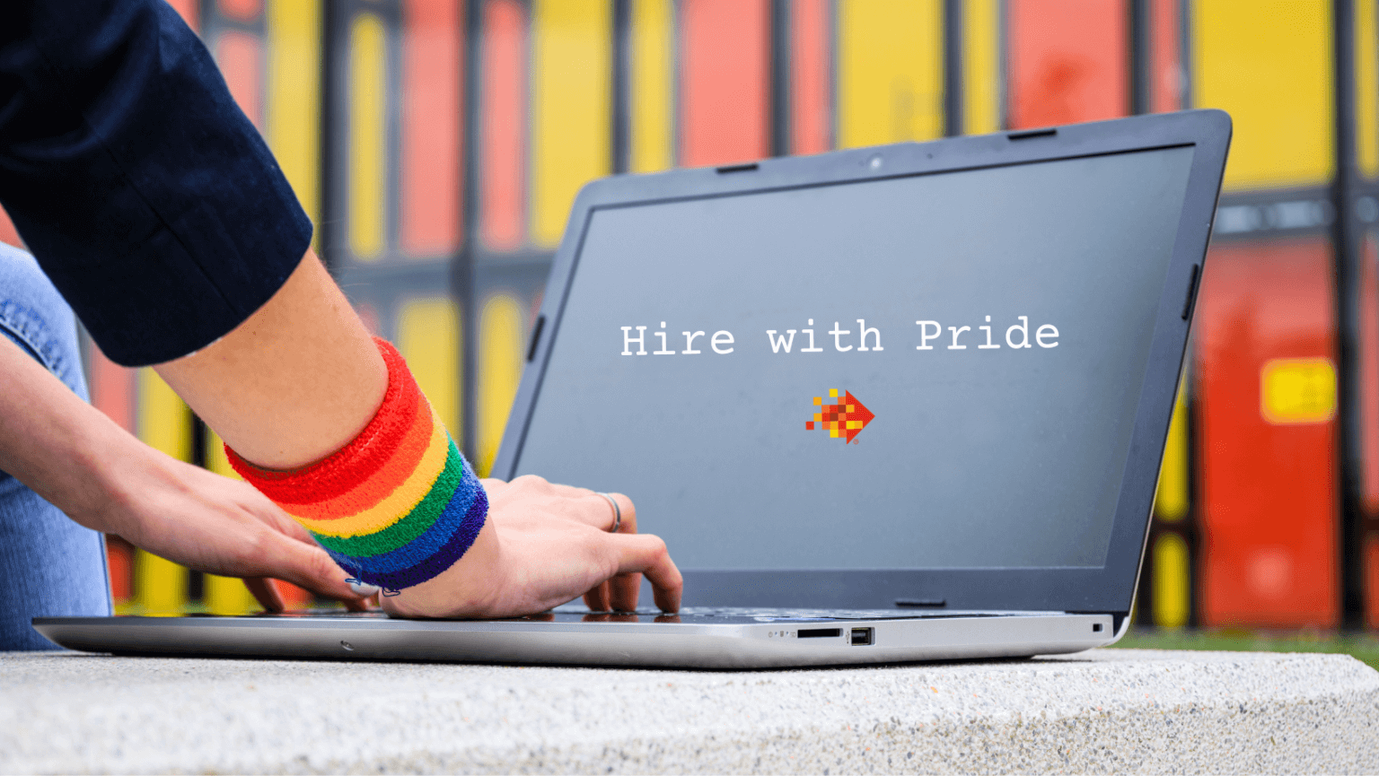 A person wearing a rainbow wristband types on a laptop. The screen displays the text "Hire with Pride" along with a pixelated arrow pointing to the right. The background consists of blurred, multicolored panels.