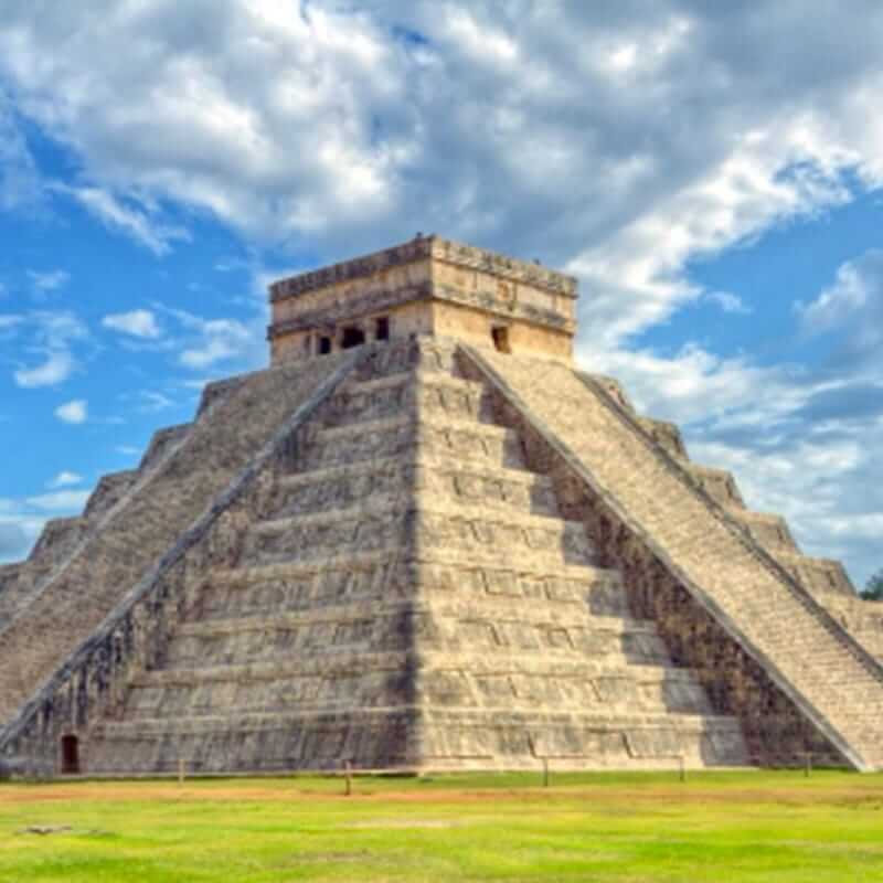 The image shows the Pyramid of Kukulcán, also known as El Castillo, at the Chichen Itza archaeological site in Mexico. The pyramid is a large step pyramid with four sides, each having 91 steps leading to a single temple at the top. The sky is partly cloudy.