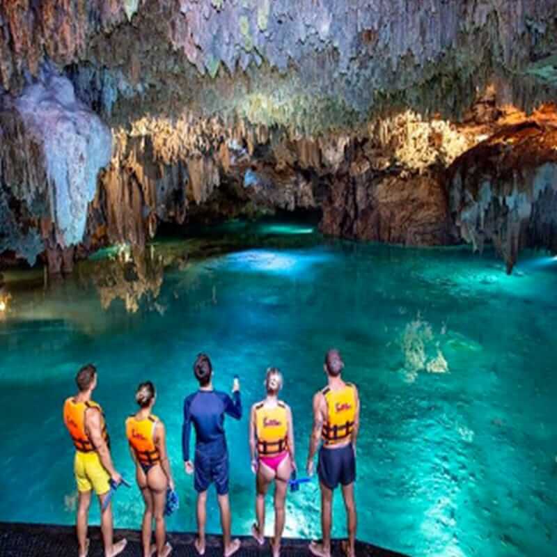 A group of five people wearing swimsuits and life jackets stand on the edge of a cave pool, admiring the bright turquoise water and stalactites hanging from the ceiling. The cave is illuminated, highlighting the natural rock formations.