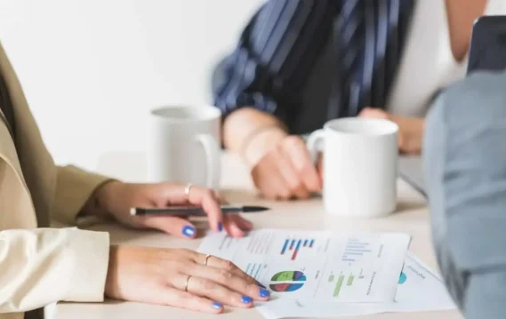 10-Skills-blog-header Two professionals discuss documents with graphs and charts on a table, accompanied by coffee mugs and a laptop in a bright, modern workspace.