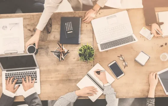 Aerial view of a collaborative workspace with hands writing, laptops, notebooks, and coffee cups on a wooden table.