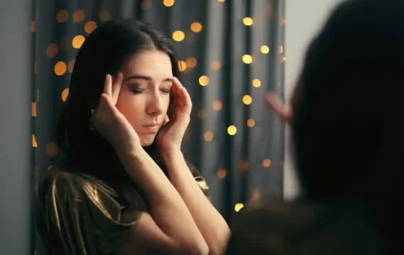 A woman stands in front of a mirror with her eyes closed and fingers on her temples, expressing signs of stress or burnout