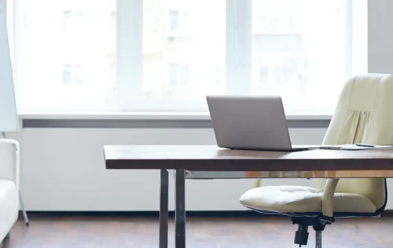 Modern minimalist office with a laptop on wooden desk, cream chair, indoor plant, and natural light from large windows.