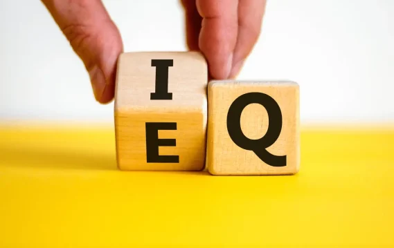Wooden blocks displaying "IEQ" and "EQ" with a hand rotating the "I" block, symbolizing emotional vs. intellectual intelligence