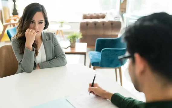 A woman in a grey blazer appears anxious during a job interview while a man holding a pen reviews her resume.