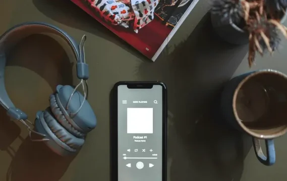 Flat lay of a smartphone playing a podcast, with headphones, coffee mug, and magazine on a table.