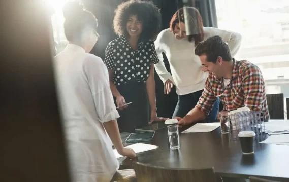 Coworkers smiling and collaborating around a table in a sunlit office meeting room.