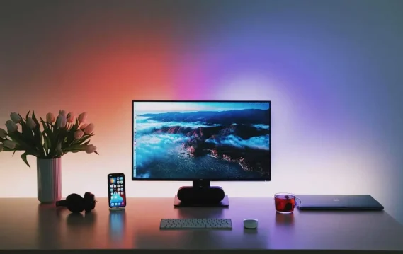 Modern workspace with a monitor showing a scenic landscape, a smartphone, laptop, cup of tea, and vase of tulips, highlighting a productive and calming environment.
