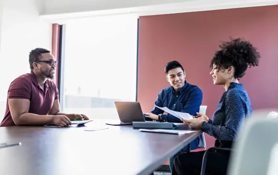 A diverse group of professionals engaged in a collaborative discussion around a conference table, reviewing documents and using a laptop.