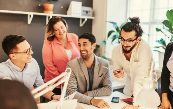 A group of five people engaged in a collaborative work session around a desk, surrounded by plants and office supplies.